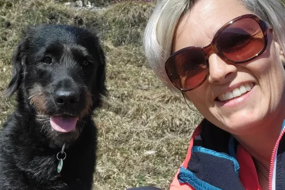 Frau mit Sonnenbrille lächelt neben einem schwarzen Hund auf einer Wiese.