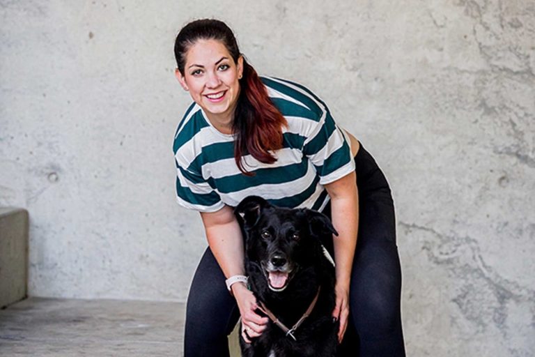 Eine lächelnde Frau kniet neben einem schwarzen Hund in einem modernen Raum.