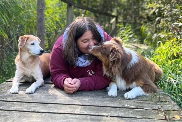 Interview mit Vanessa über Maulkörbe im Training Frau liegt auf einem Holzsteg und wird von zwei Hunden liebevoll angeleckt.